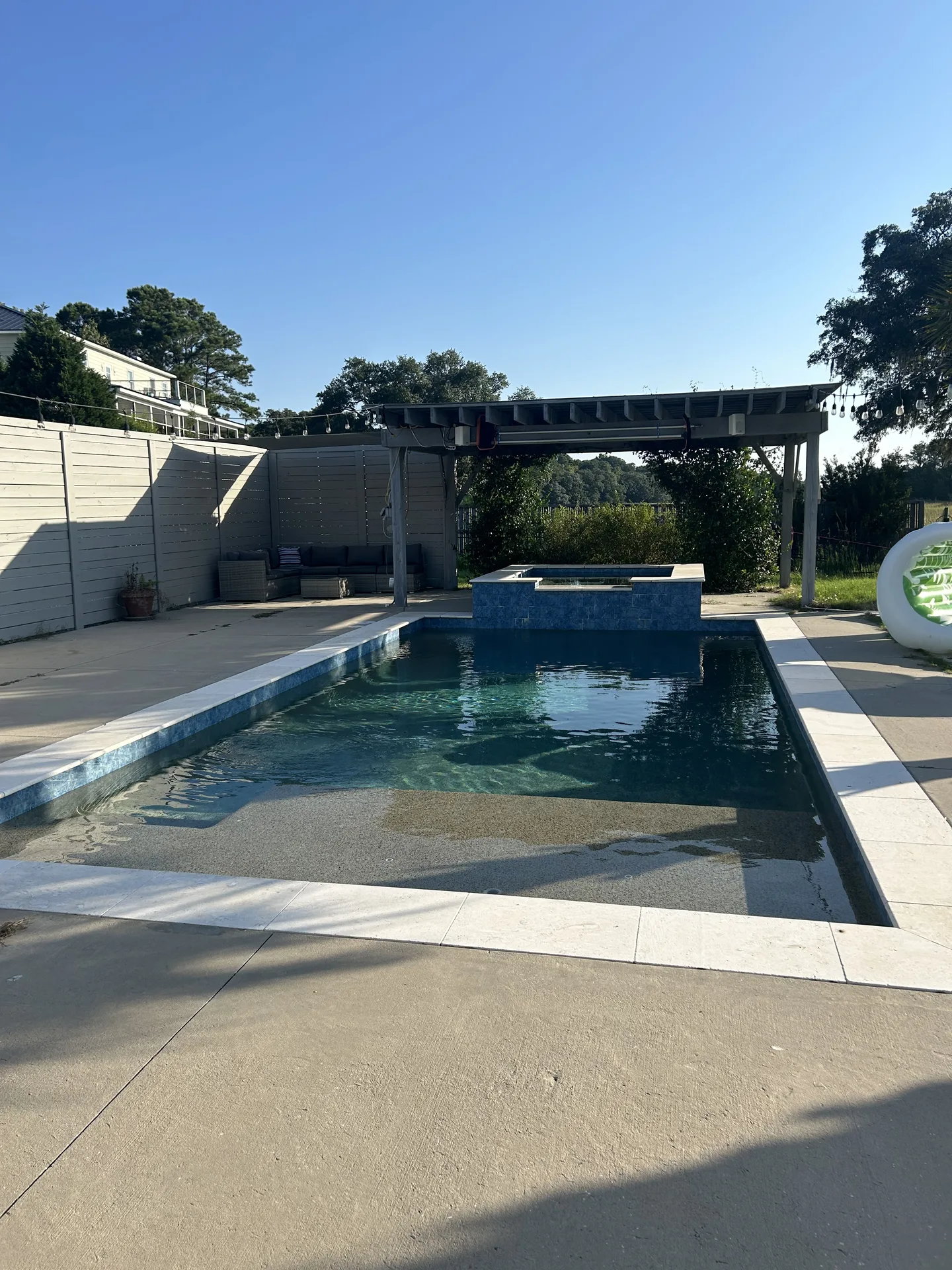 Saltwater pool with raised spa viewed from the pergola — lounge chairs, string lights, and palm trees framing turquoise water