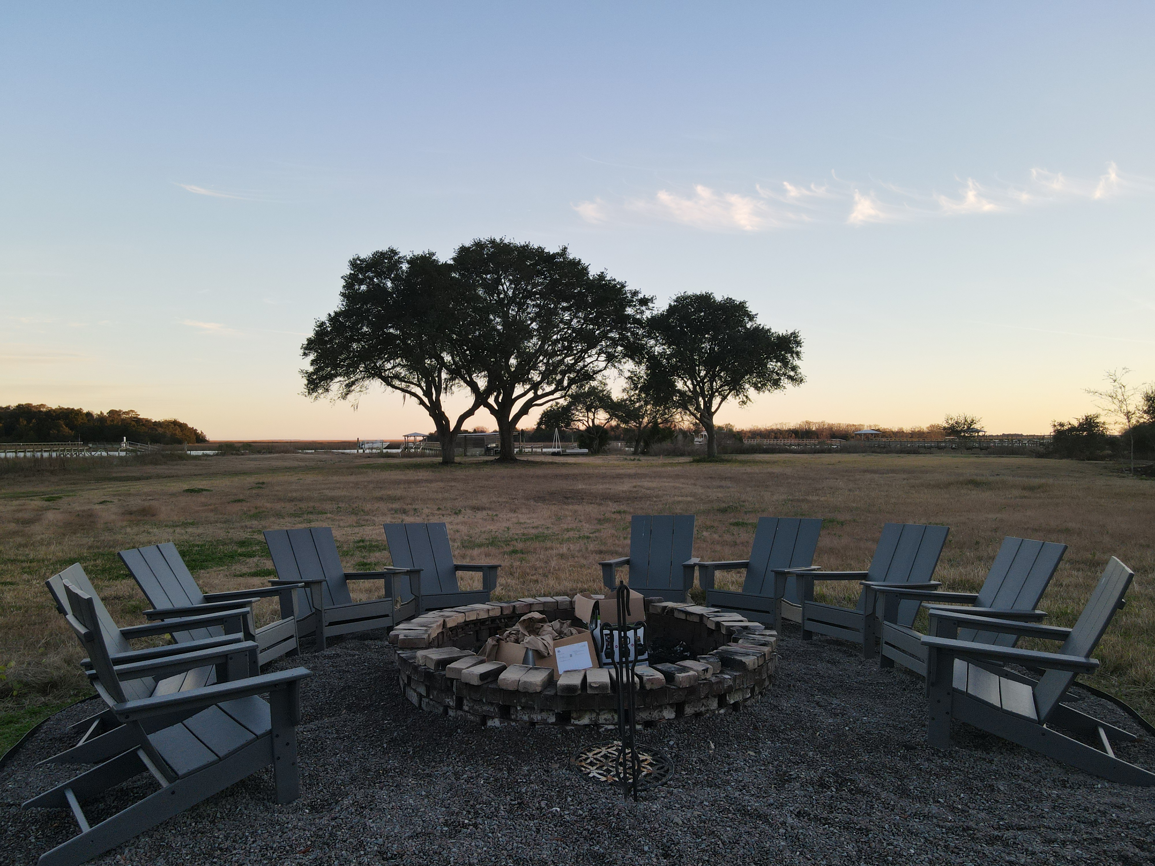 Stone fire pit ringed by Adirondack chairs under ancient spreading live oaks with the waterway visible at sunset — the evening gathering spot